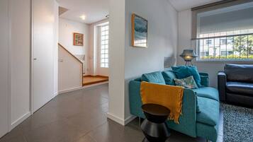 A modern living room with a teal sofa, a black coffee table, and a black leather couch. There is a staircase leading to another room, and a window with a view of the outside.