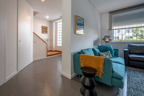 Ein modernes Wohnzimmer mit einem türkisfarbenen Sofa, einem schwarzen Couchtisch und einem schwarzen Ledersofa. Eine Treppe führt in einen anderen Raum, und ein Fenster bietet einen Blick ins Freie.