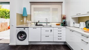 A compact kitchen with a built-in washer, a sink, and a window with a view of greenery. - Clementina: Serene Garden and Pool Stay w/Parking (Lisboa)