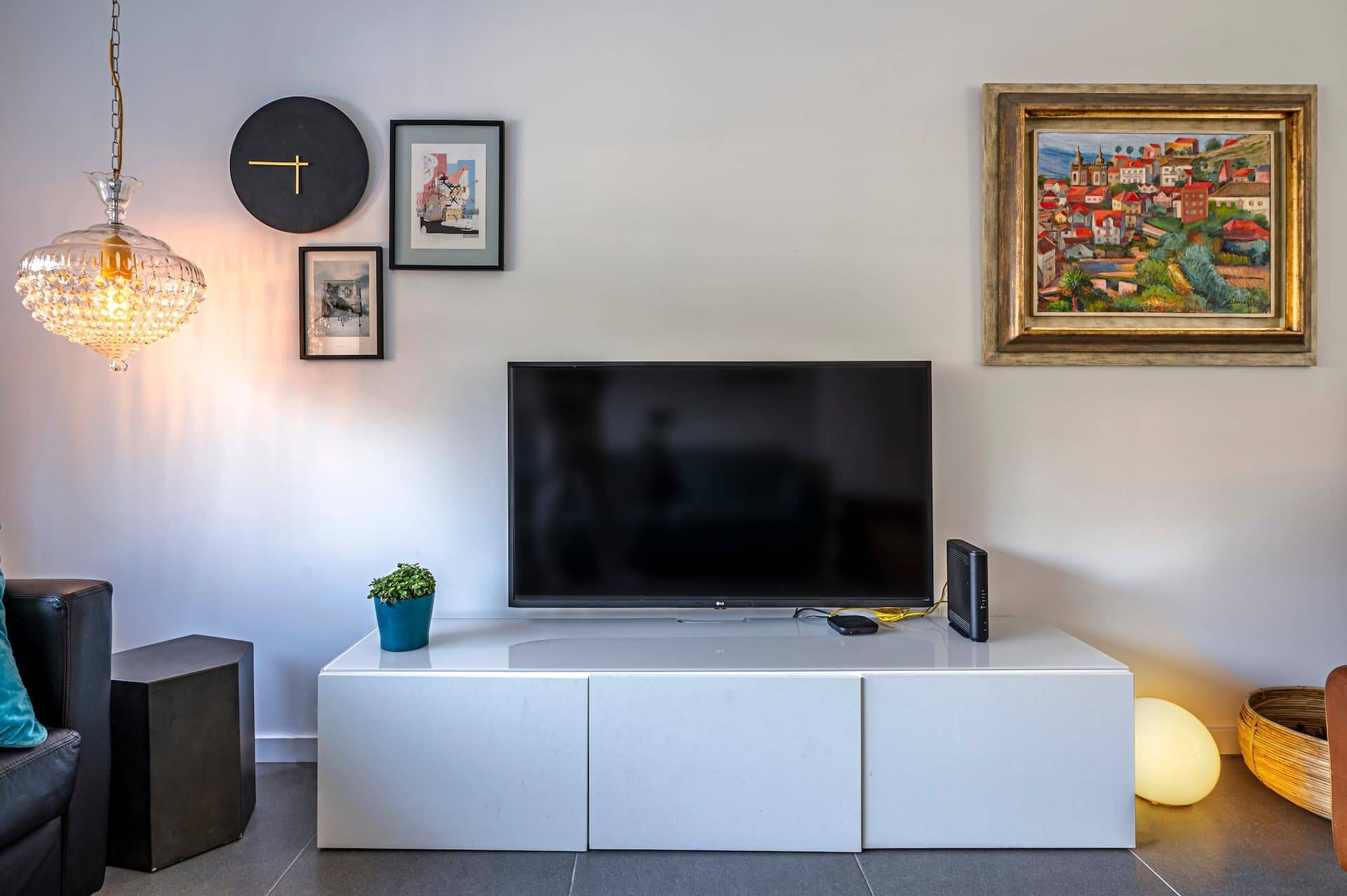 A modern living room with a flat-screen TV on a white cabinet, a clock, framed artwork, and a hanging light fixture.