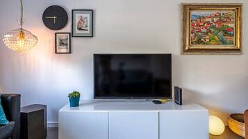 A modern living room with a flat-screen TV on a white cabinet, a clock, framed artwork, and a hanging light fixture.