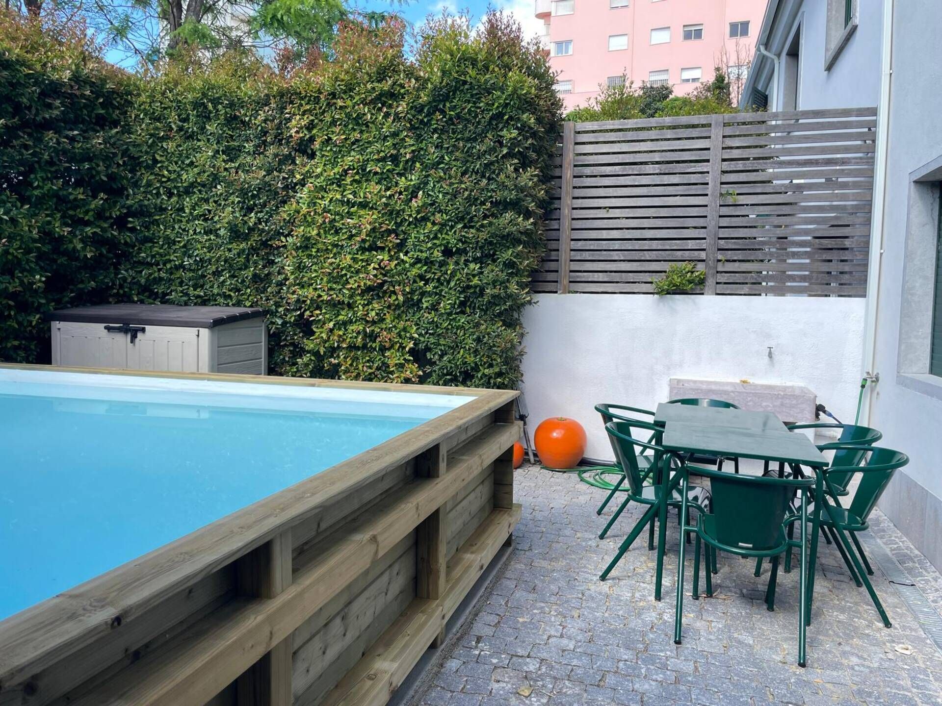 A swimming pool with a wooden deck, green outdoor furniture, and a hedge in the background.