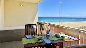 Een houten tafel met een groene tafelkleed, omringd door stoelen en diverse decoratieve objecten op een balkon met uitzicht op een strand.
