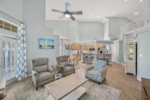 A spacious living area with a high ceiling, ceiling fan, and a view of the kitchen and dining area.
