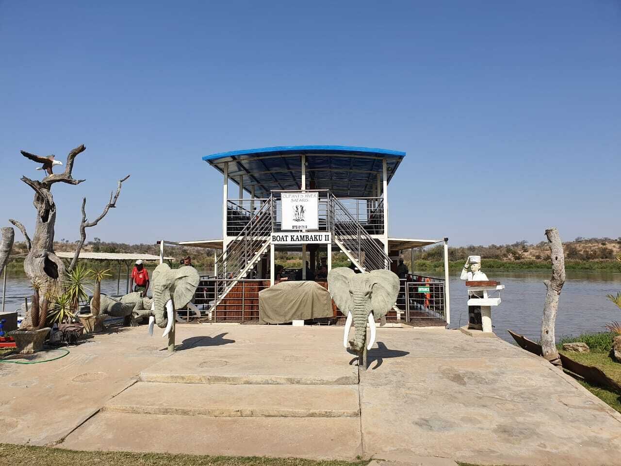 A boat named 'Boat Kambaku II' docked at a riverside location with a covered shelter and a statue of a man.