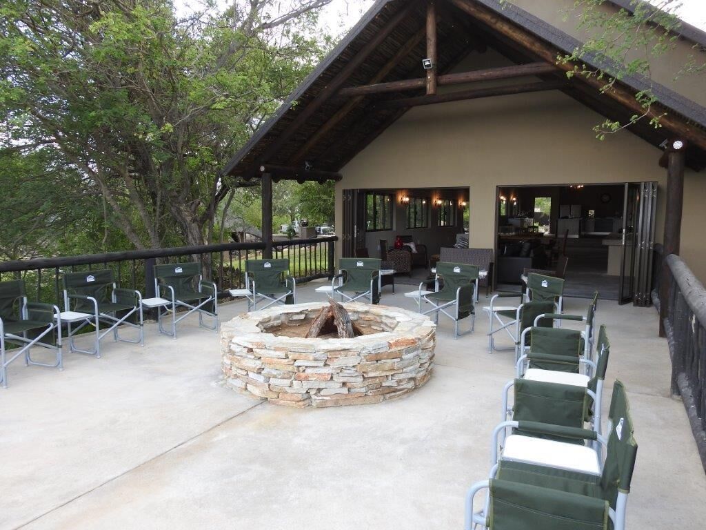An outdoor seating area with stone fire pit and green chairs, a covered patio, and a building with a wooden roof.