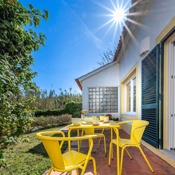 A patio with yellow chairs and a table, a white building with a window, and a clear blue sky.