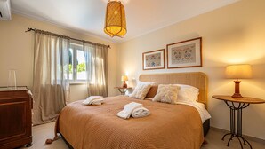 Ein ordentlich bezogenes Bett mit Handtüchern, ein Nachttisch mit Lampe, ein Fenster mit Vorhängen und zwei gerahmte Bilder an der Wand.