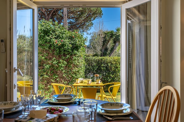 An open patio with yellow chairs and a table set for dining, overlooking a garden with various plants and trees.