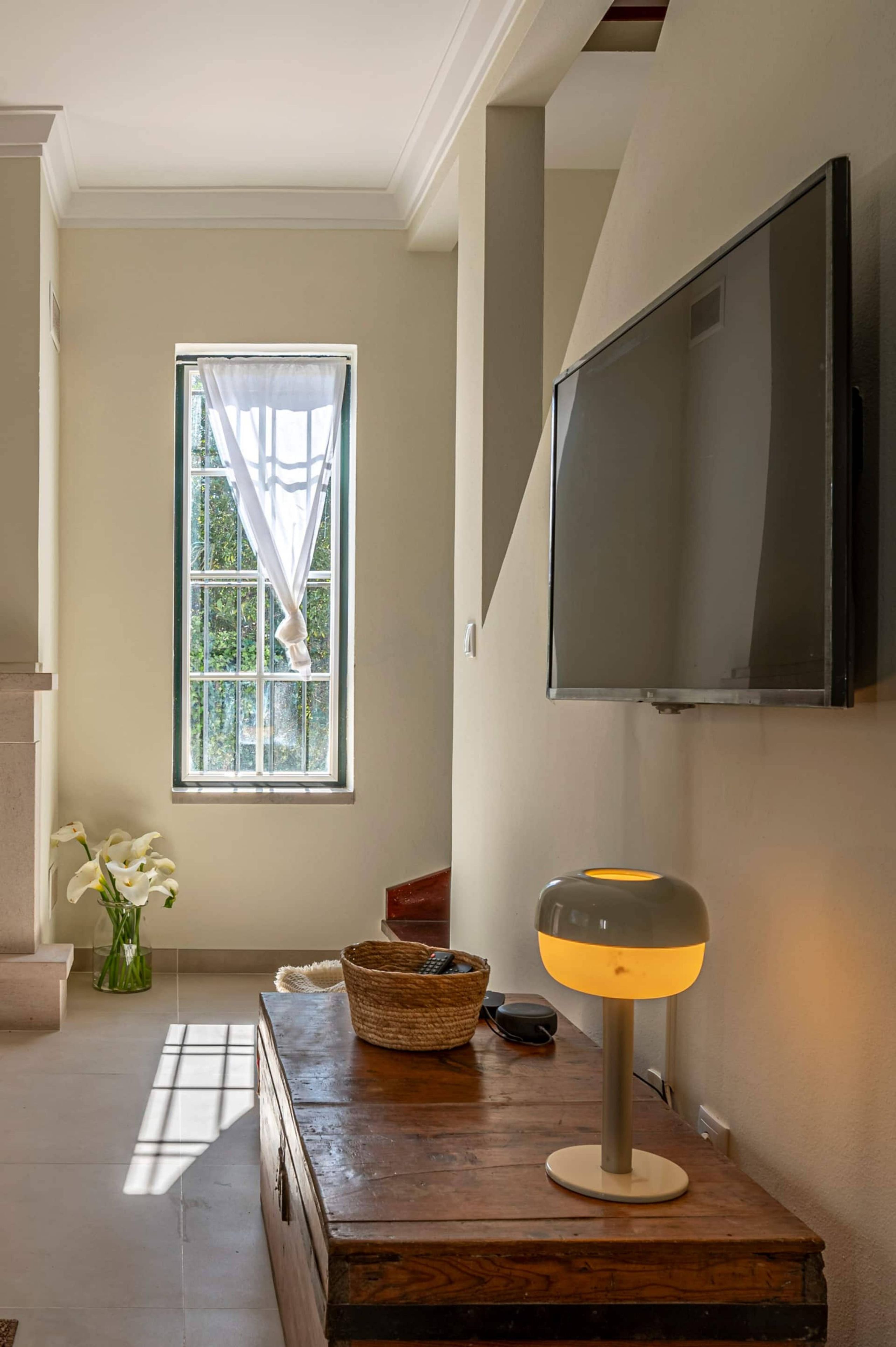 Une pièce dotée d’une grande fenêtre, d’un téléviseur à écran plat fixé au mur, d’une table en bois avec une lampe et d’un vase rempli de fleurs.