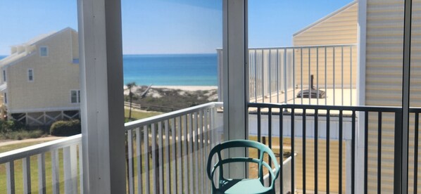 A balcony with a view of the ocean and beach, a chair, and a building with a white exterior.
