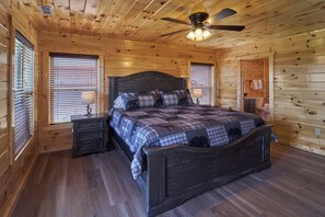 Dormitorio de cabaña de madera con cama, mesitas de noche, ventilador de techo y ventana con persianas.