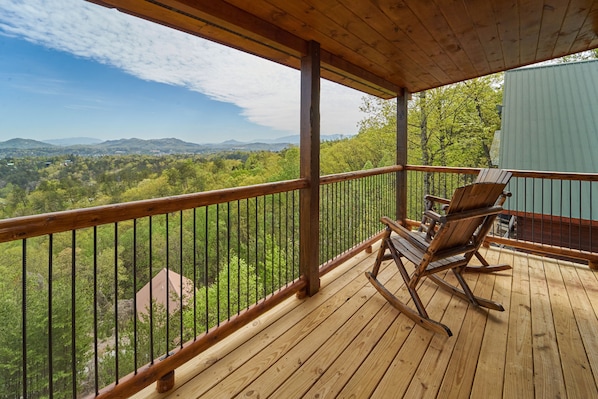 A wooden deck with a railing, two rocking chairs, and a view of a forested landscape with mountains in the distance.