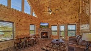 A log cabin interior with a fireplace, wooden furniture, and large windows. - Cabin w/Breathtaking Views, Hot Tub & Game Room (Sevierville)