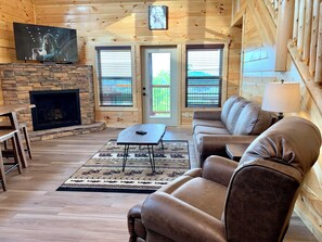 A cozy living room with a stone fireplace, leather sofa, and a flat-screen TV mounted on the wall. - Cabin w/Breathtaking Views, Hot Tub & Game Room (Sevierville)