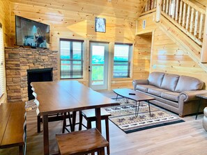 A wooden interior with a stone fireplace, a sofa, a dining table, and a staircase.