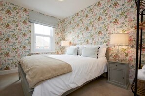Une chambre à coucher avec un papier peint à motifs floraux, un lit avec une literie blanche, deux lampes de chevet et une fenêtre avec des stores.