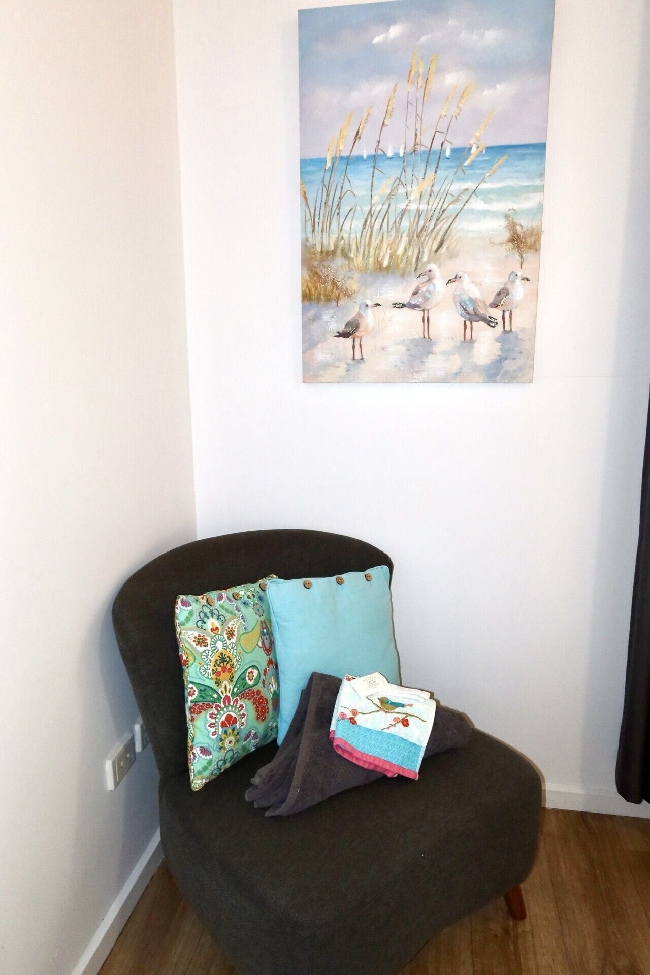 Un coin de pièce avec un fauteuil de couleur marron foncé, un coussin turquoise et un coussin à motifs floraux colorés. Un tableau représentant des mouettes et des roseaux est accroché au mur.