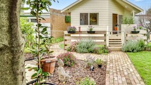 Une maison de plain-pied avec un porche à l’avant, un chemin en briques et un jardin agrémenté de diverses plantes et fleurs.