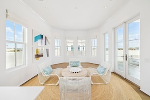 A bright room with white walls, large windows, wicker furniture, and a ceiling fan.