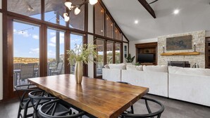 A spacious living area with a wooden dining table, large windows, and a stone fireplace. - The Sanctuary at Canyon Lake | 3 Bed, 3 Bath (Canyon Lake)