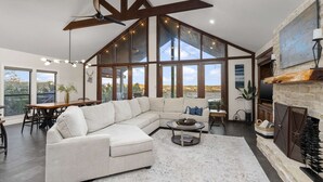 A spacious living room with a large L-shaped sectional sofa, a fireplace, and large windows offering a view of the outdoors. - The Sanctuary at Canyon Lake | 3 Bed, 3 Bath (Canyon Lake)