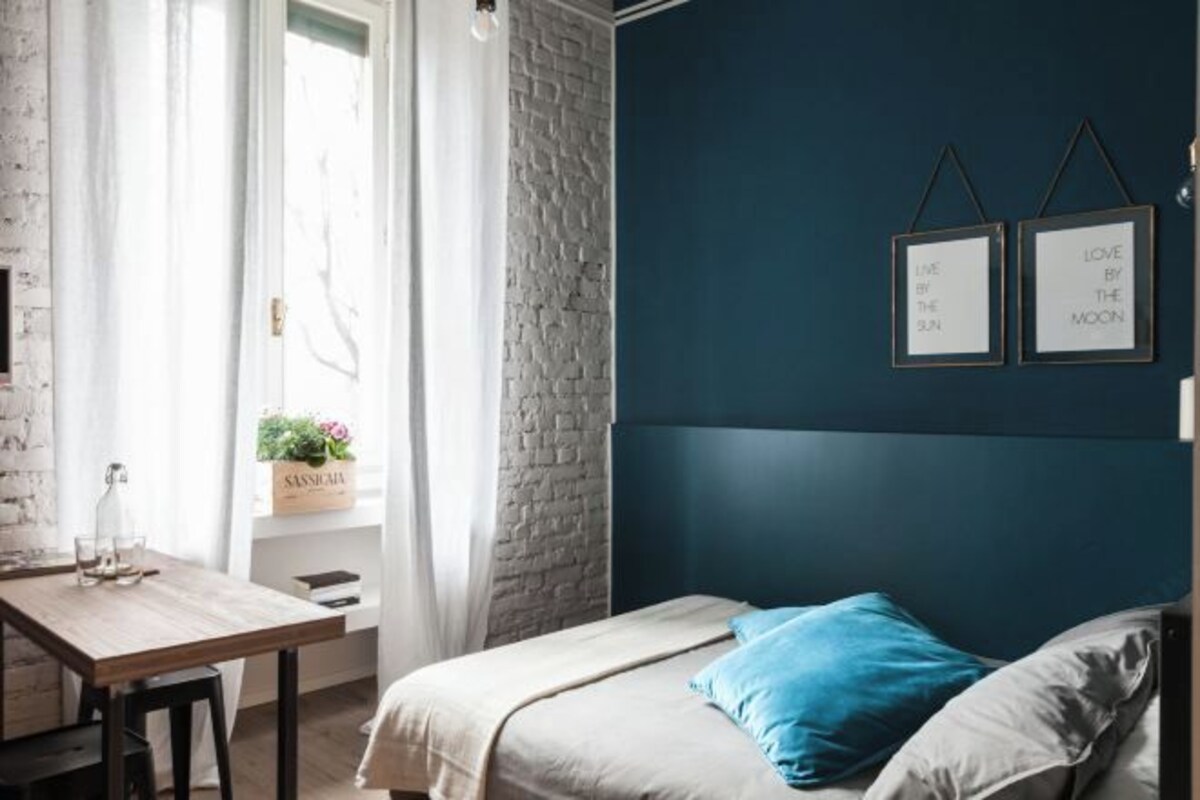 Un dormitorio con cama, escritorio y ventana con cortinas. Hay dos cuadros en la pared.