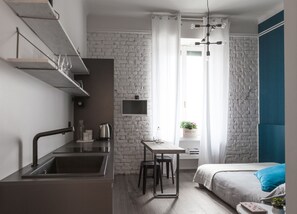 A modern bedroom with a bed, bedside table, and a small desk with a chair. There is a kitchen area with a sink and a kettle. - Ca Bela Lambrate (Milan)