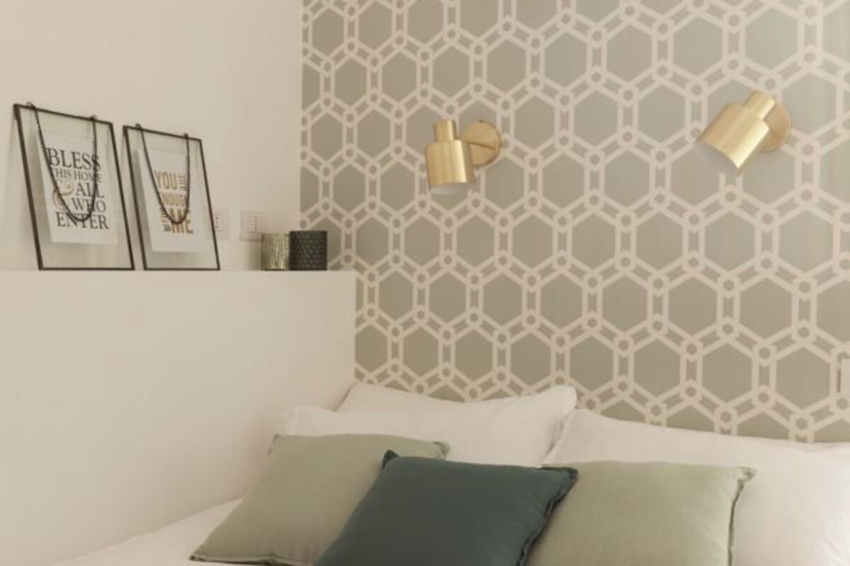 Un dormitorio con papel tapiz estampado geométricamente, una cama blanca con muchos almohadones y dos lámparas de pared.