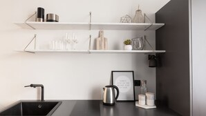 Una cocina moderna con un lavamanos negro, un hervidor de acero inoxidable y estantes blancos con cristalería y utensilios de cocina.