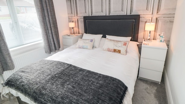 Cama bem arrumada com cabeceira preta, roupa de cama branca e manta cinza. Duas mesas de cabeceira com luminárias e um criado-mudo também com uma luminária. O quarto tem uma parede estampada e uma janela com cortinas.