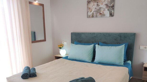 A neatly made bed with a beige duvet and two blue pillows, a bedside table with a lamp, and a wall-mounted mirror. - Depie Luxury Apartment in Pastida Rhodes (Rhodes)