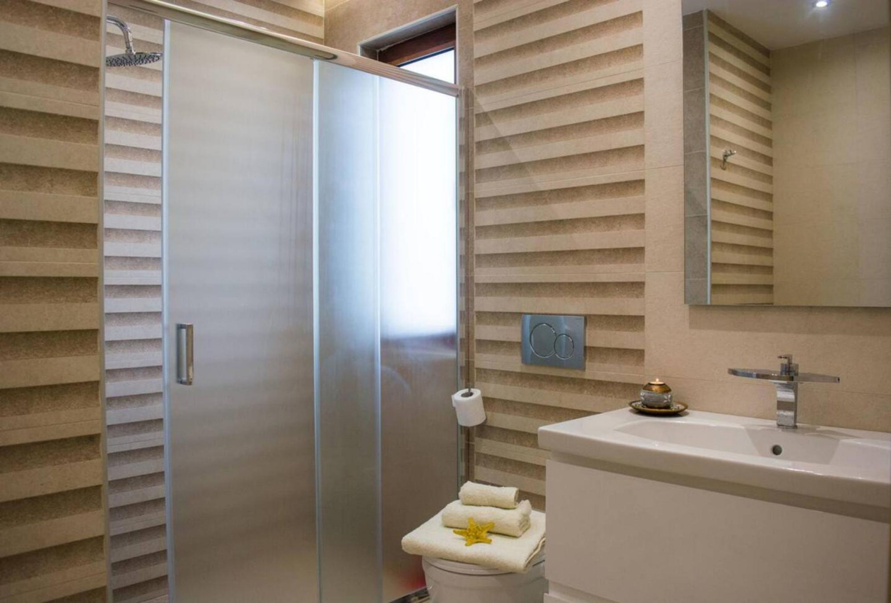 Un baño moderno con mampara de vidrio, inodoro con toalla y decoración de estrellas de mar, y lavamanos con un objeto dorado en la encimera.