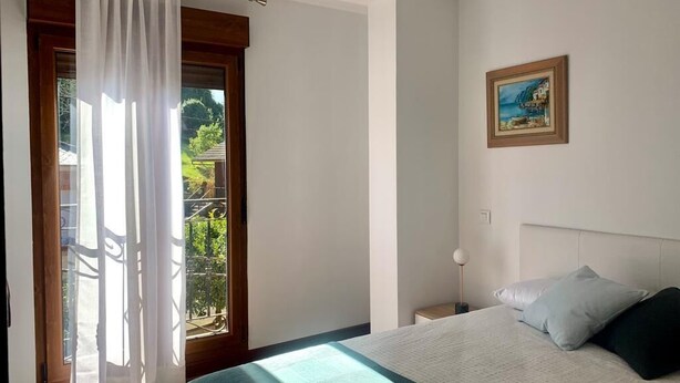 A bedroom with a bed, a nightstand, a painting, and a view through a window with curtains.