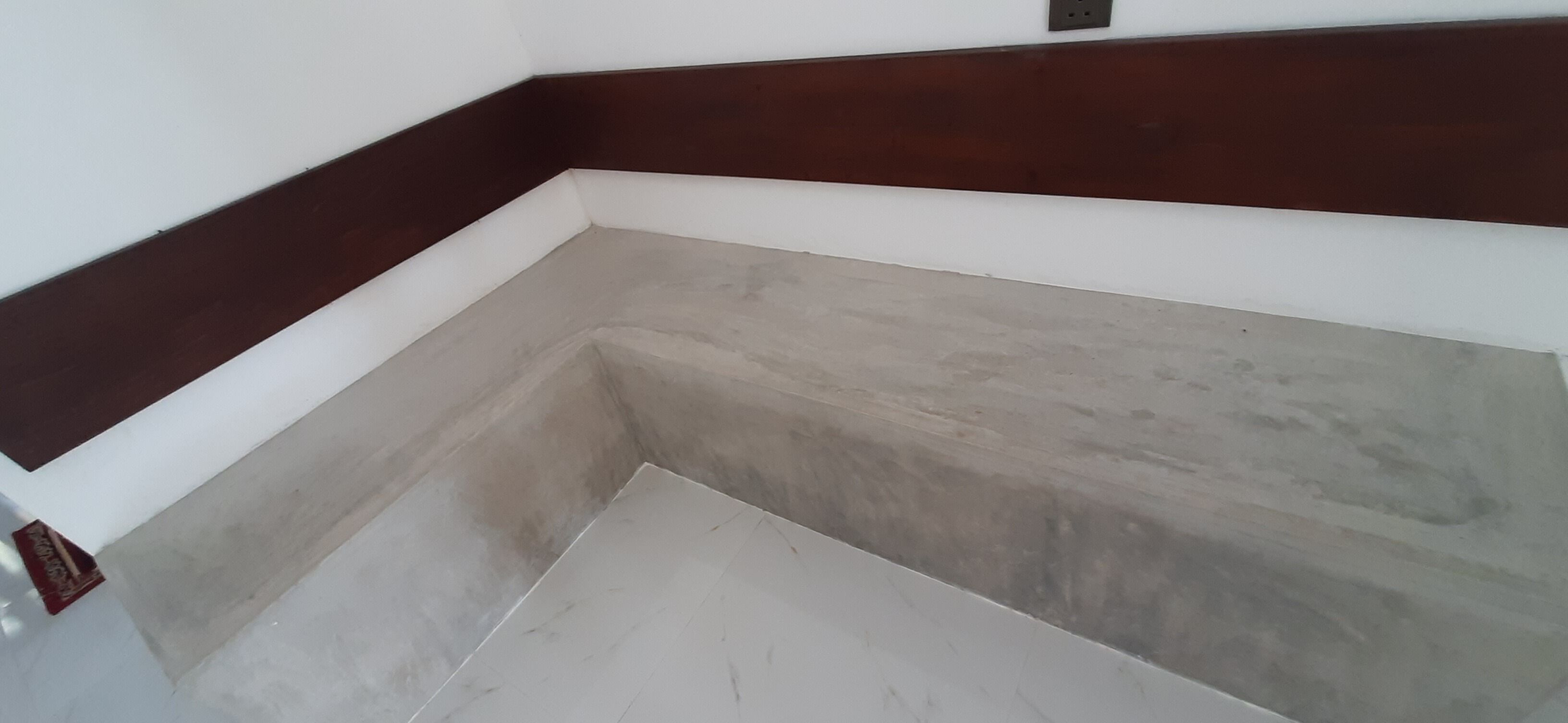 A corner of a room with a white wall and a dark brown baseboard.