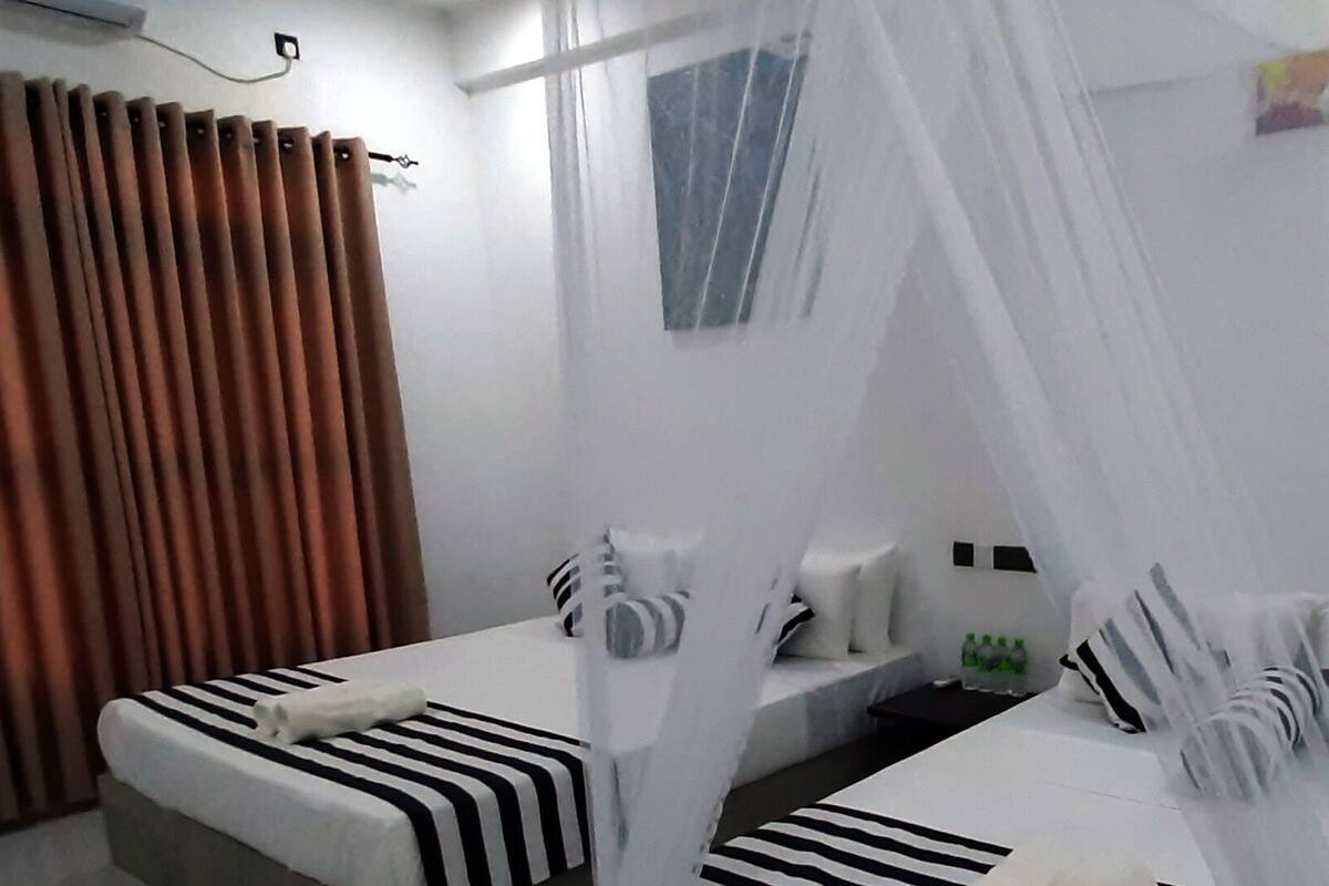 A hotel room with two beds, a window with curtains, a bedside table with toiletries, and a wall-mounted shelf.