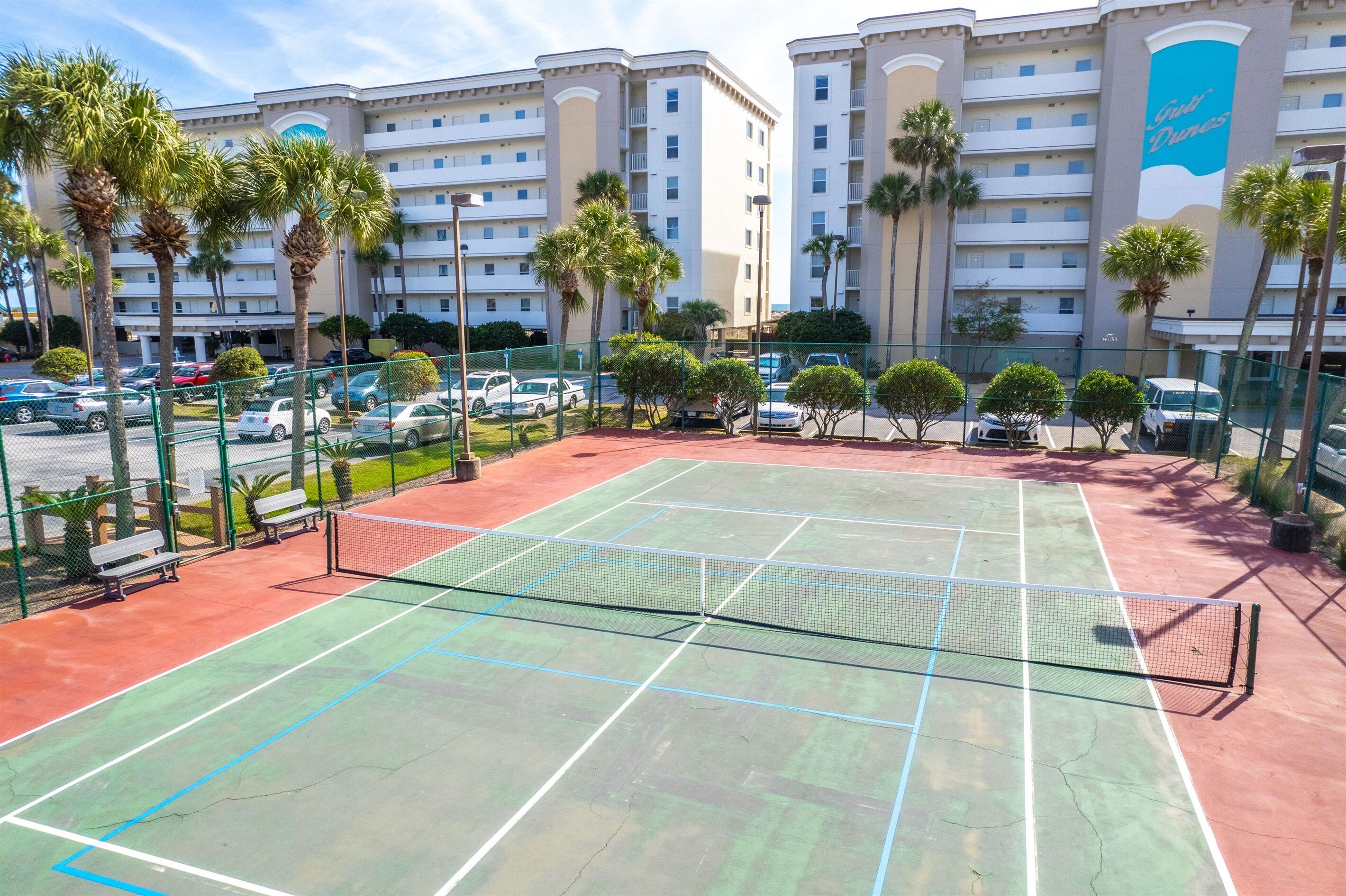A tennis court with a net, surrounded by a fence, palm trees, and a hotel building in the background.
