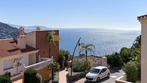 Un quartier résidentiel en bord de mer, avec une voiture blanche garée dans une allée, une vue dégagée sur la mer et un bâtiment doté d’un toit en tuiles.