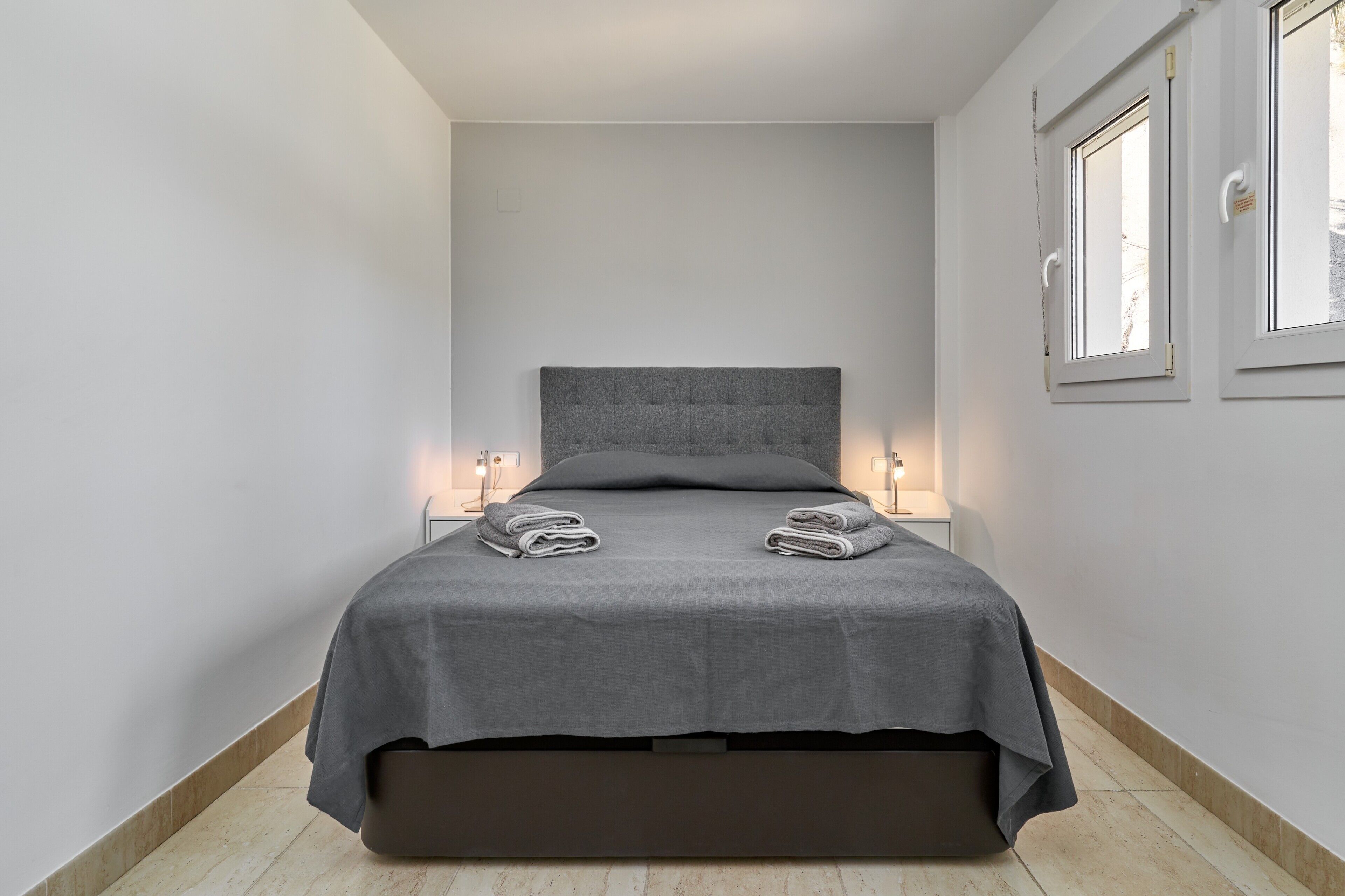 A bed with a grey headboard, two bedside tables with lamps, and a window on the right wall.
