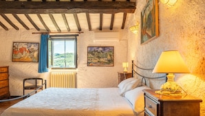 Un dormitorio con cama, cómoda de madera, mesita de noche con lámpara, ventana con vistas y dos cuadros en la pared.