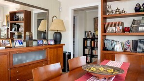 A dining area with a wooden table, chairs, a red and white patterned table runner, a decorative bowl, and a bookshelf with books and decorative items. - Millenium by Interhome (Premià de Dalt)