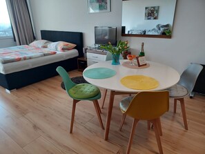 A modern bedroom with a bed, a TV, a dining table with chairs, and a small plant.