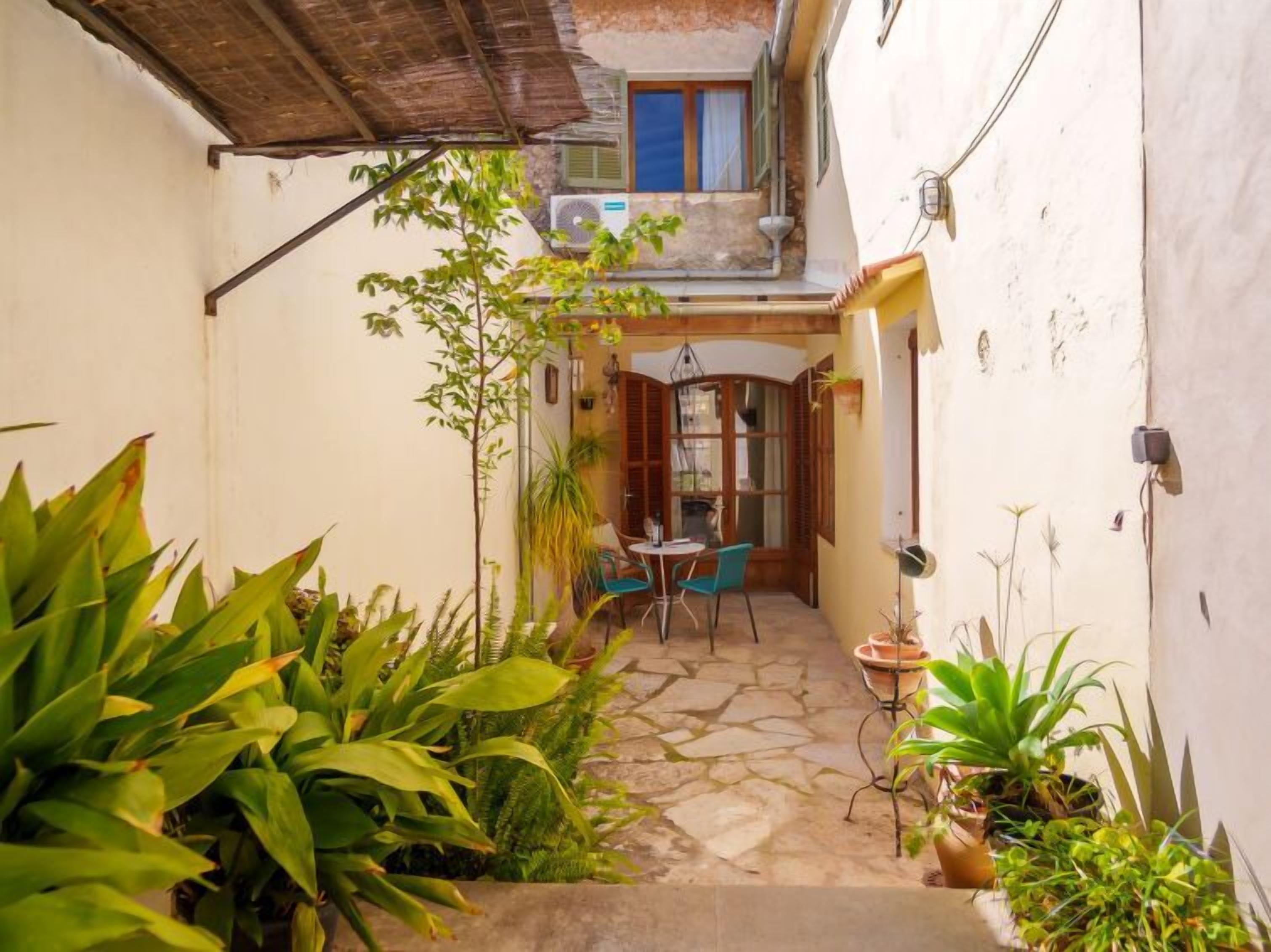 A narrow courtyard with a stone floor, a small table with chairs, and various potted plants.