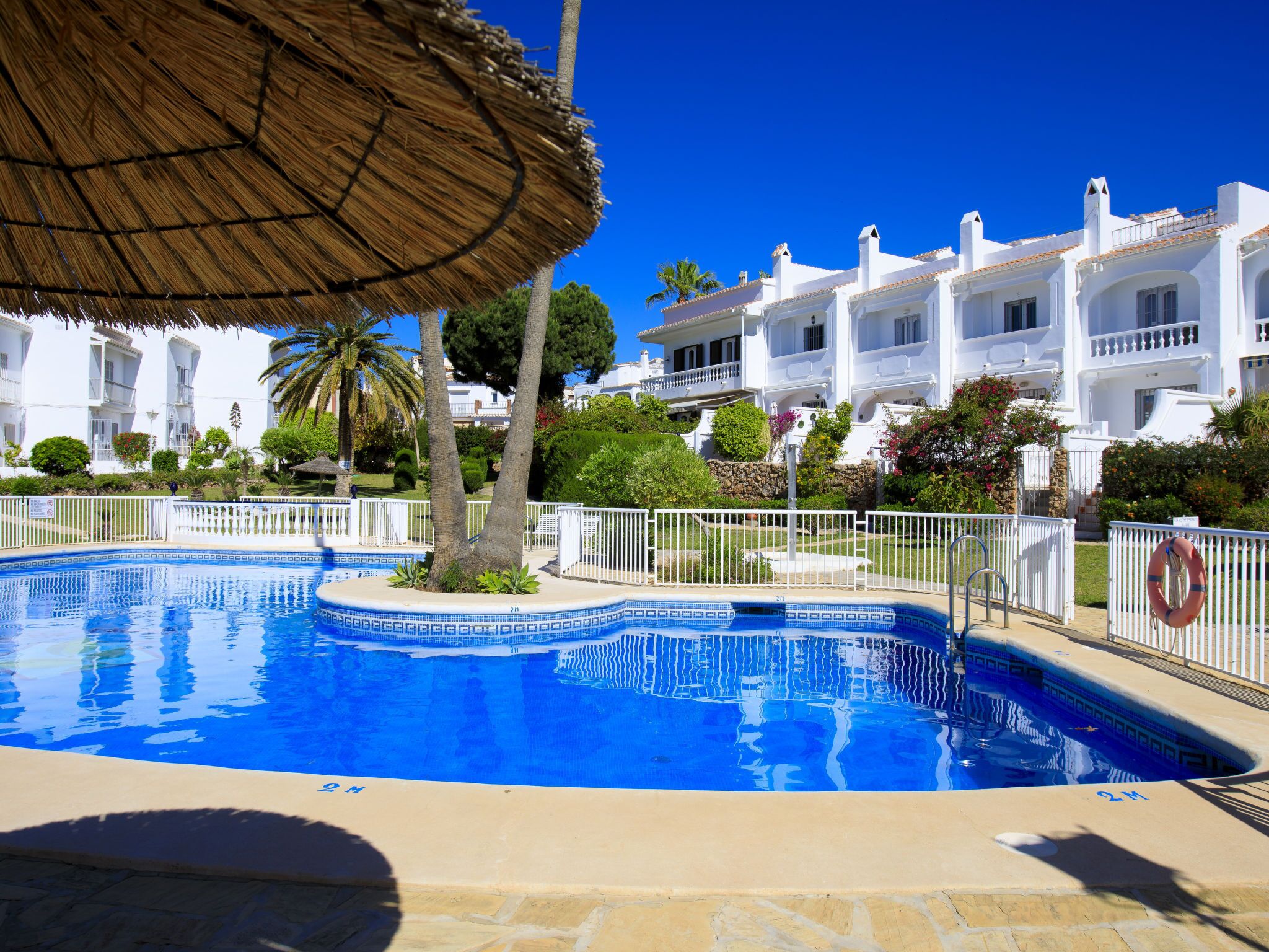 Une piscine dont le bord est recouvert de carreaux, entourée d’une clôture blanche, et un parasol en chaume situé à proximité.