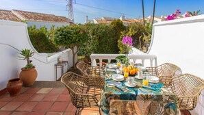 A terrace with a dining table set for breakfast, surrounded by wicker chairs, potted plants, and a view of the neighborhood. - Jardines de Nerja by Interhome (Nerja)