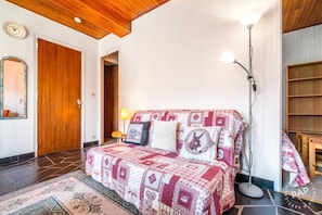 Ein Schlafzimmer mit Holzbalkendecke, einem Bett mit gemusterten Betttextilien, einer Nachttischlampe und einer Wanduhr.