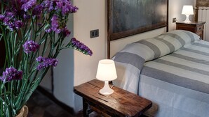 Une chambre à coucher avec un lit, une table de chevet en bois sur laquelle se trouve une lampe, un grand miroir et un vase rempli de fleurs violettes.
