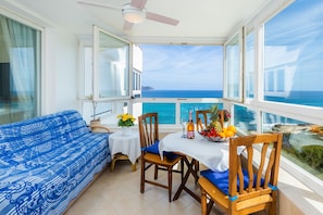 Un balcon avec vue sur la mer, un canapé bleu à motifs, des chaises en bois, une table avec une bouteille et un panier de fruits.