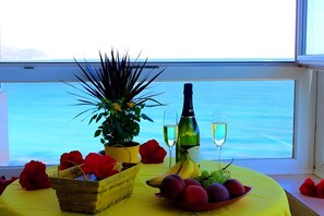 Une table avec des fruits, une bouteille et des verres, placée près d’une fenêtre donnant sur la mer.