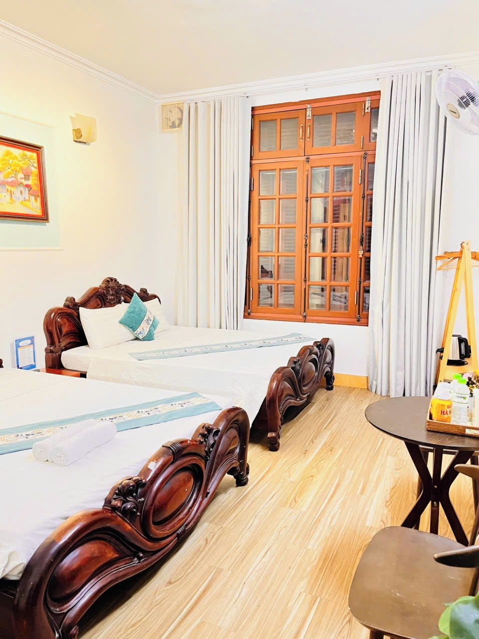 A hotel room with a double bed, a wooden floor, a small table with a chair, and a window with curtains.
